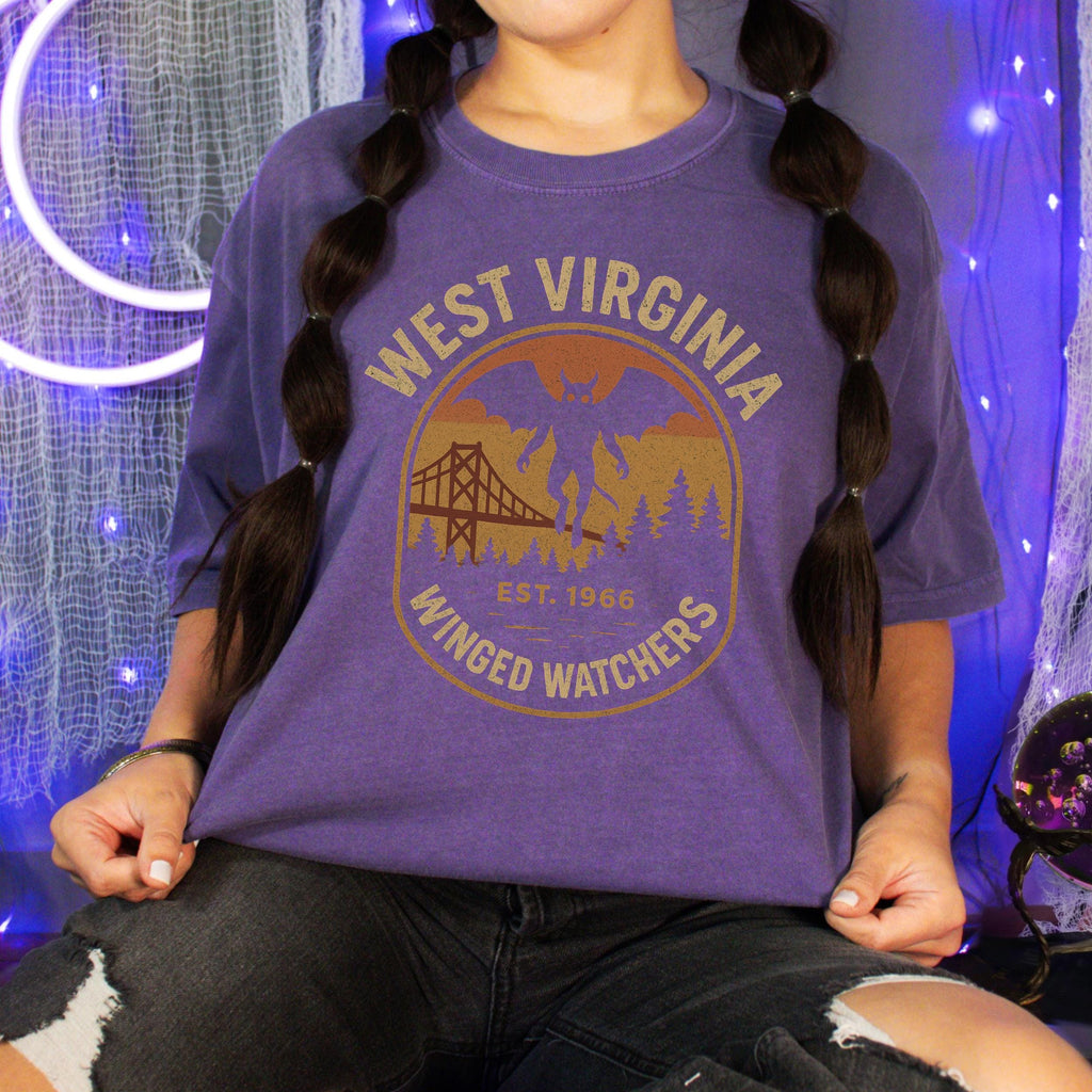 The Faithful Familiar purple Comfort Colors t-shirt featuring a vintage Mothman graphic with a bridge and forest.